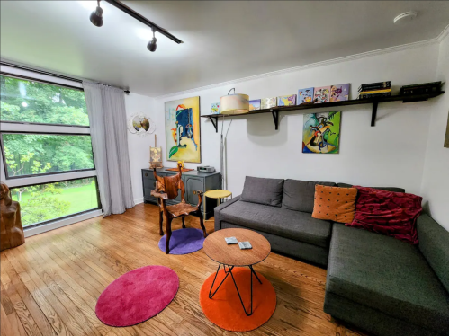 Cozy living room with a gray sectional sofa, colorful rugs, artwork on the walls, and large windows overlooking greenery.