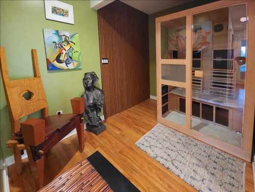 A cozy room featuring a wooden chair, a sculpture, a sauna, and colorful artwork on the walls.