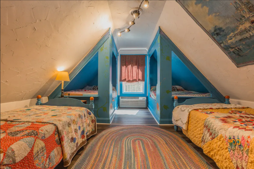 Cozy attic bedroom with two twin beds, colorful quilts, and sloped ceilings, featuring a rug and window with curtains.
