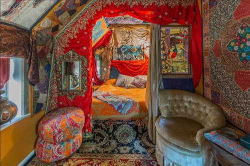 A vibrant, bohemian-style bedroom with red drapes, colorful textiles, and ornate decor, featuring a cozy bed and seating area.