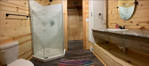 Cozy bathroom with wooden walls, a glass shower, a rustic sink, and a colorful rug on the floor.