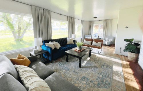 Cozy living room with large windows, comfortable seating, and a coffee table, surrounded by greenery outside.