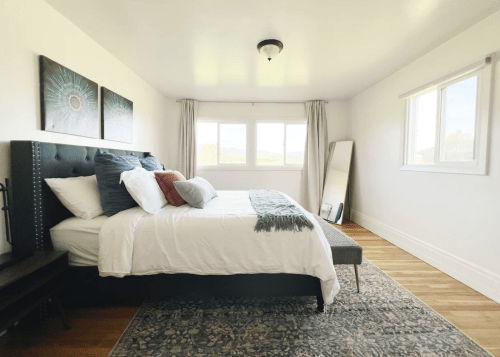 Bright bedroom with a large bed, decorative pillows, a mirror, and windows offering natural light.
