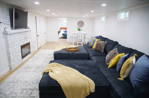 Cozy basement living area with a large sectional sofa, decorative pillows, a TV, and a dining space in the background.