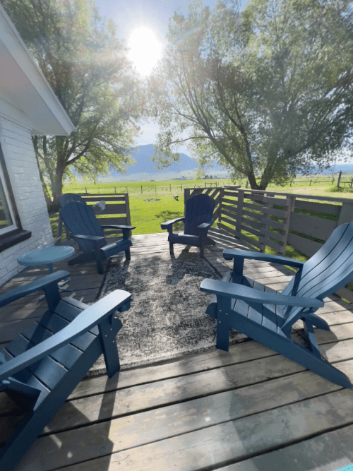 A sunny outdoor patio with blue chairs and a rug, surrounded by greenery and distant mountains.