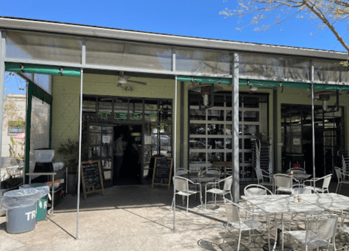 A modern café with outdoor seating, large windows, and a green awning on a sunny day.