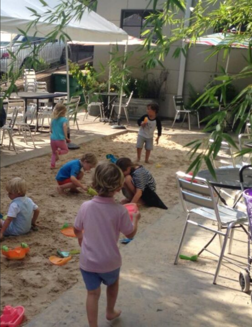 Children playing in a sandy area with toys, surrounded by tables and chairs under shaded canopies.