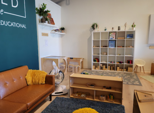 A cozy educational playroom with a brown couch, shelves, toys, and a blue rug. Bright and inviting atmosphere.