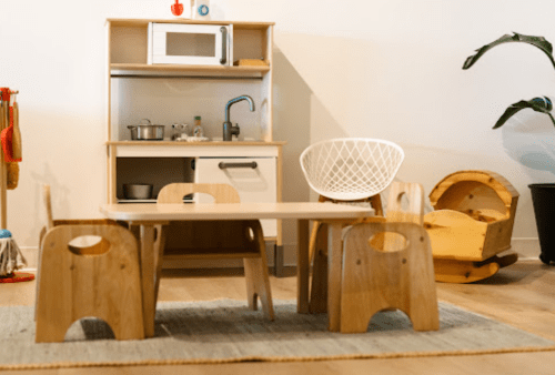 A cozy playroom with a wooden table, chairs, a toy kitchen, and a rocking chair on a rug.