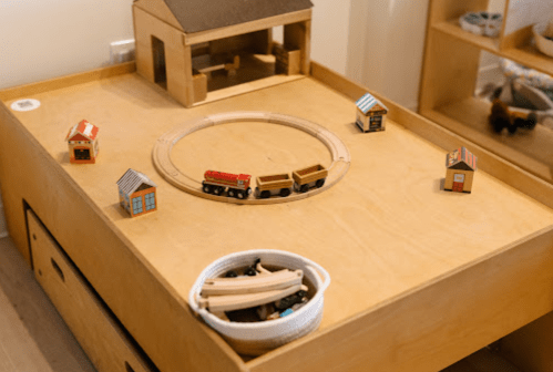 A wooden play table with a train track, toy houses, and a basket of small wooden toys.