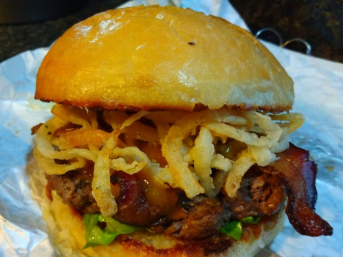 A delicious burger with lettuce, bacon, crispy onion rings, and a golden bun, served on a crumpled wrapper.