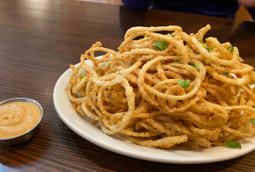 A plate of crispy onion rings piled high, served with a small bowl of dipping sauce on the side.