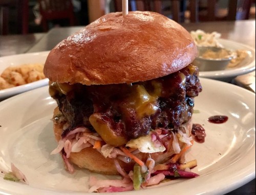 A close-up of a juicy burger with melted cheese and coleslaw on a toasted bun, served on a white plate.