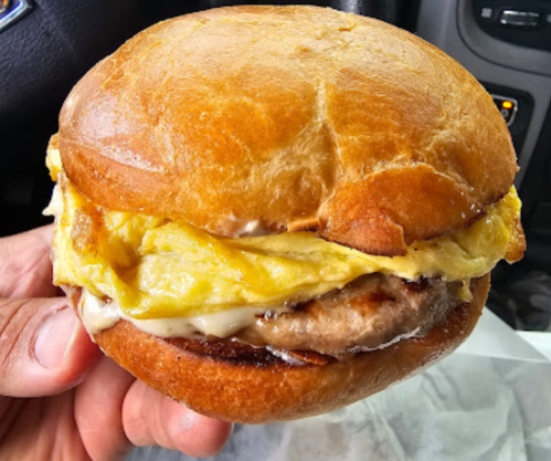 A hand holding a breakfast sandwich with a fluffy egg, sausage patty, and creamy sauce on a toasted bun.