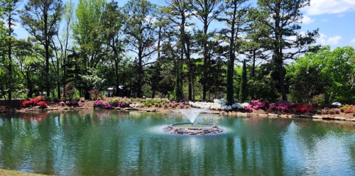 A serene pond surrounded by colorful flowers and trees, featuring a fountain in the center under a clear blue sky.