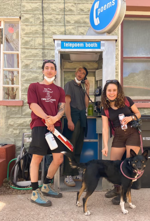 Three people stand by a telepoem booth, smiling. A dog sits in front, and one person is on the phone.
