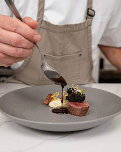 A chef drizzles sauce over a gourmet dish featuring meat, caviar, and garnishes on a gray plate.