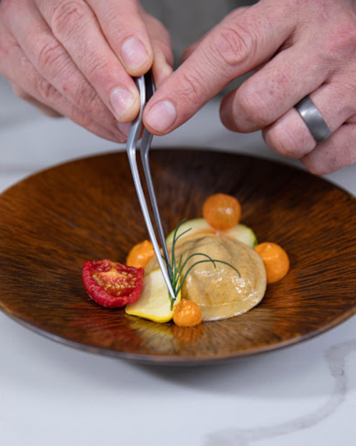 A person uses tweezers to arrange colorful garnishes on a gourmet dish in a brown bowl.