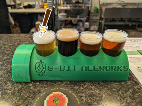 Four small glasses of beer in a green holder labeled "8-BIT ALEWORKS" on a countertop.