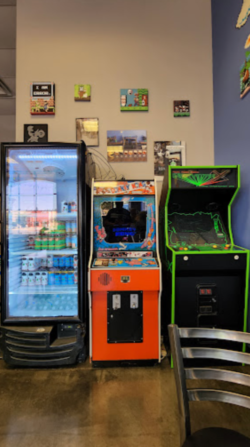 A colorful arcade game cabinet next to a fridge and another arcade machine in a casual dining space.