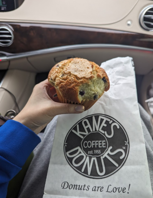 A hand holding a blueberry muffin next to a Kane's Donuts bag inside a car.
