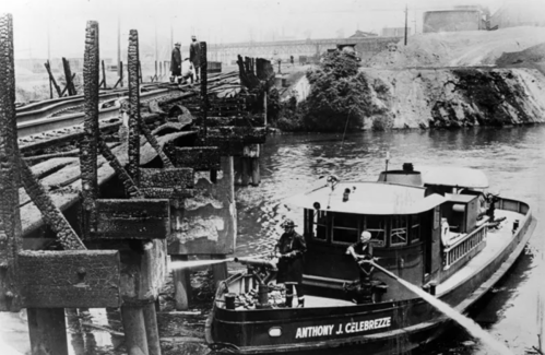 A historic black-and-white photo of a boat near a dilapidated bridge, with workers on the structure and water below.