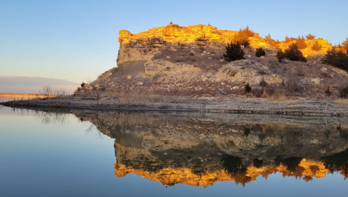 A rocky cliff bathed in golden light reflects in calm water at sunset, surrounded by sparse vegetation.