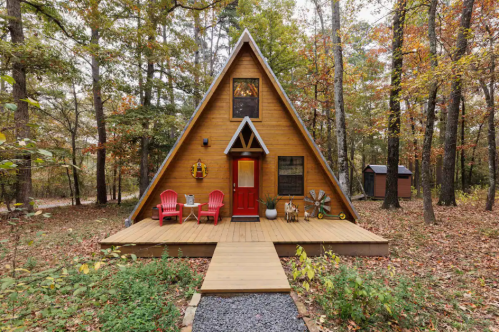 A cozy A-frame cabin surrounded by trees, featuring a red door and two red chairs on a wooden porch.