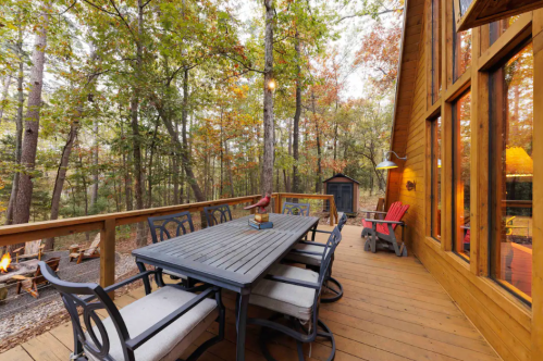 A wooden deck with a dining table and chairs, surrounded by trees in autumn colors, with a cozy cabin in the background.