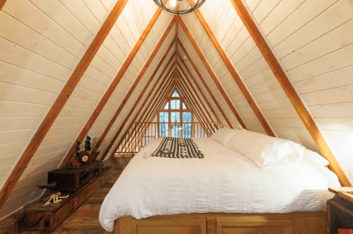 Cozy A-frame bedroom with wooden beams, a large window, and a white bedspread, creating a warm, inviting atmosphere.