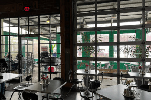 Interior of a cafe with large glass doors, green accents, and tables set for dining. Outdoor seating visible.