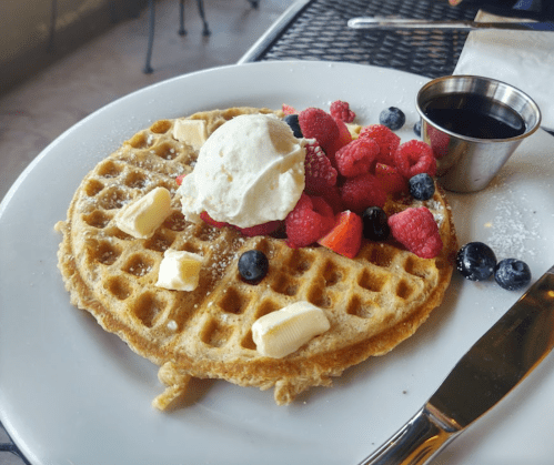 A golden waffle topped with fresh berries, whipped cream, butter, and a small cup of syrup on a white plate.