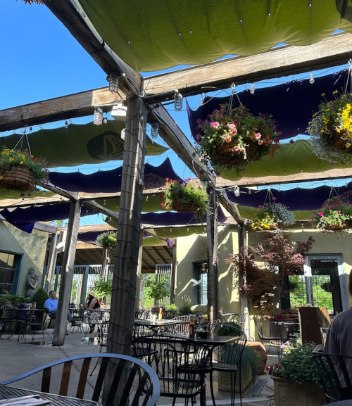 Outdoor dining area with hanging flower baskets and colorful canopies, featuring tables and chairs under a clear blue sky.