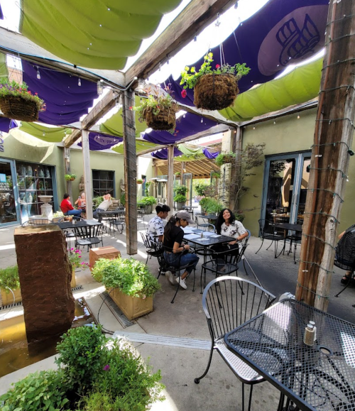 Outdoor dining area with black tables and chairs, surrounded by greenery and colorful canopies. People are enjoying their meals.