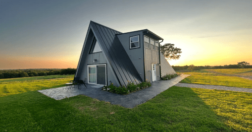 A modern A-frame house with a sloped roof, surrounded by green grass and a sunset sky in the background.