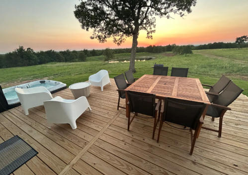 A wooden deck with a dining table and chairs, overlooking a sunset and green landscape, with a hot tub nearby.