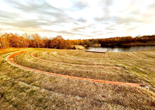 A scenic view of a grassy amphitheater overlooking a calm pond, surrounded by trees under a cloudy sky.