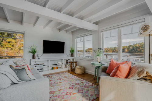 Cozy living room with large windows, a TV, and colorful cushions, featuring a warm, inviting atmosphere.