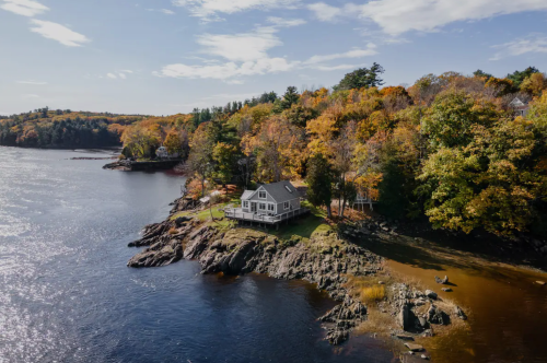 A scenic view of a house on a rocky shore, surrounded by autumn foliage and calm waters under a partly cloudy sky.