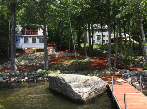 A serene lakeside scene featuring two houses among trees, a rocky shore, and a wooden dock extending into the water.