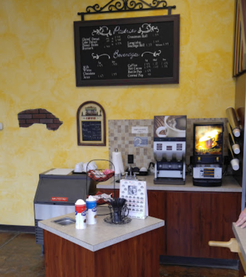 A cozy café counter with a menu board, beverage machines, and a display of desserts in a warm yellow setting.