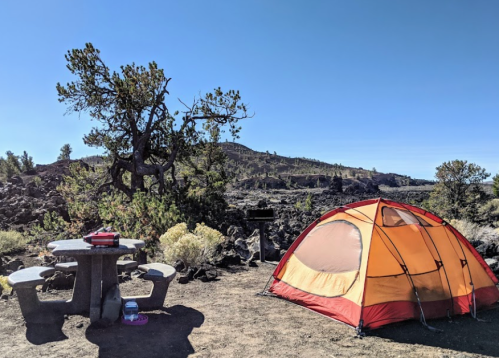 A bright orange tent set up at a campsite with a picnic table and rocky landscape under a clear blue sky.