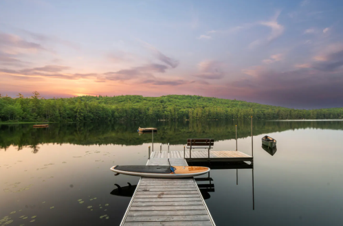 A serene lake at sunset, featuring a wooden dock, a paddleboard, and lush green hills in the background.