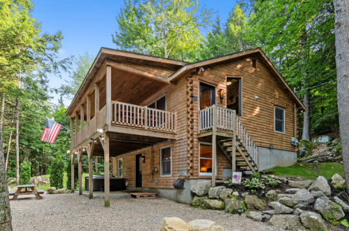 A wooden cabin surrounded by trees, featuring a porch, stairs, and an American flag, set in a gravel area.