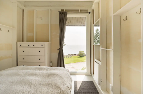 A bright bedroom with a bed and dresser, featuring a view of the sea through a large window.