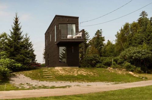 A modern black house on a grassy hill, surrounded by trees, with a balcony and large windows.