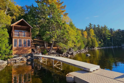A wooden cabin by a serene lake, surrounded by trees, with a dock extending into the calm water.