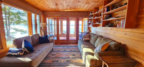 Cozy wooden living room with two sofas, a rug, and large windows overlooking a lake. Shelves filled with books.