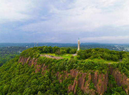 Aerial view of a green hilltop with a tower, surrounded by trees and a cloudy sky in the background.