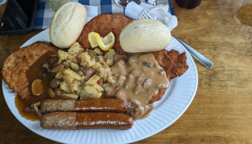 A plate of food featuring fried meat, sausage, potatoes, gravy, and two rolls, garnished with lemon slices.
